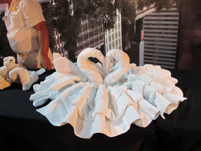 Photo shows two terry-cloth swan with their beaks together such that their necks are arched in the shape of a heart, and surrounding them are rows of white  pleated terry-cloth.